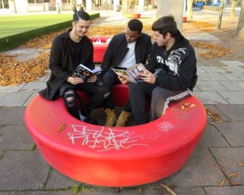 Tre tonårskillar sitter utomhus i en ring och två av dem läser ur en bok.